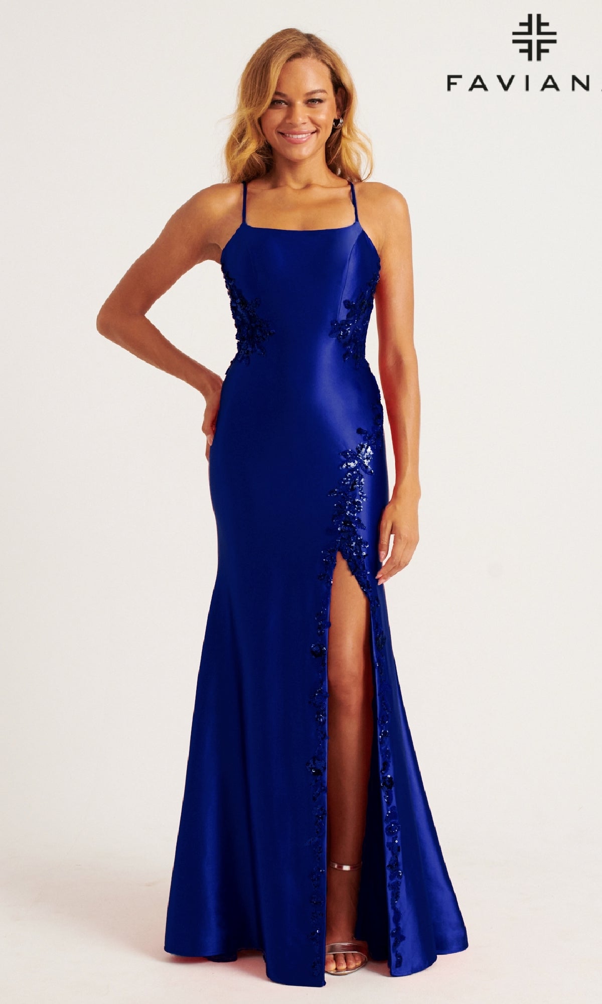 A woman wearing the sleeveless, floor-length royal blue Lace-Up Long Mermaid Prom Dress: Faviana 11205 with sequin details and a thigh-high slit stands smiling.